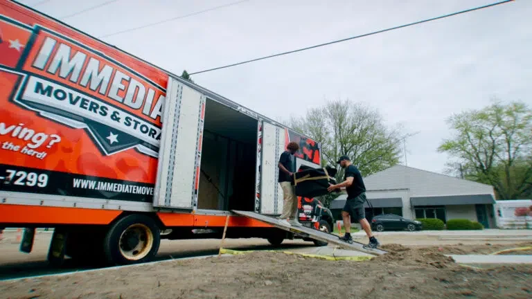 Immediate Movers project photo — two crew members walking a loaded dolly up the ramp into a branded orange Immediate Movers box truck