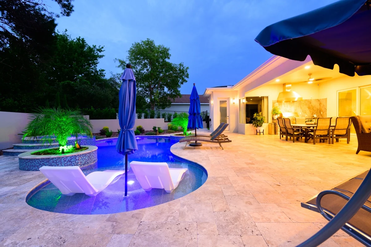 Nic’s Pool Construction project photo — a pool lit purple and green by LED at dusk, with in-water loungers, a mosaic planter and a lit outdoor kitchen behind