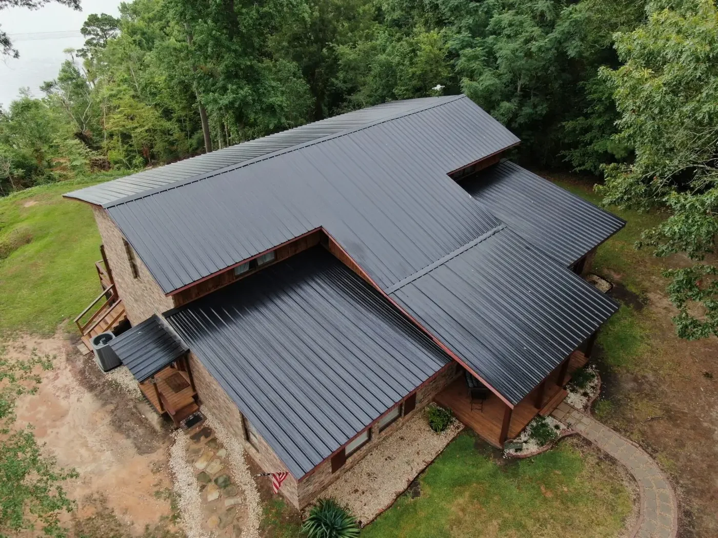 Roof Ryders project photo — a drone view of a stone and cedar lake house in East Texas under a large dark standing-seam metal roof